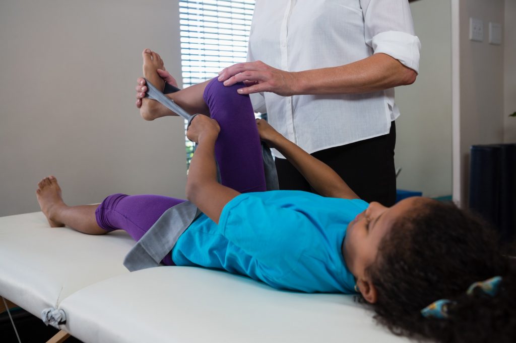 Physiotherapist assisting girl patient in performing stretching exercise from resistance band